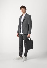 Blazer e pantaloni grigi, camicia bianca e valigetta nera. Le scarpe sono bianche con accenti sottili. Trame morbide e vestibilità su misura.