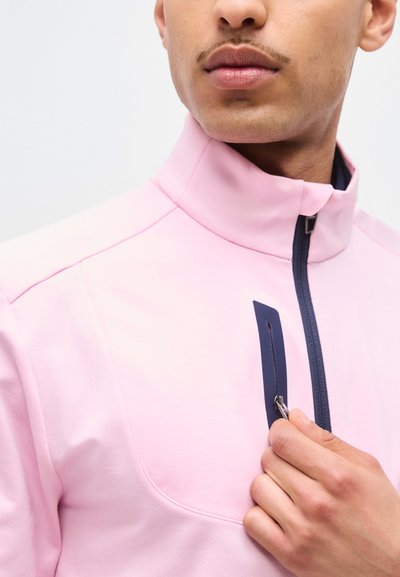 Mężczyzna w jasnoróżowej kurtce z wysokim kołnierzem, trzymający rękę na zamku kieszeni na piersi z granatowym obszyciem.