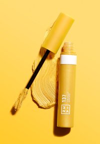 Gelbe Mascara-Tube mit mattem Finish, ausgestattet mit einem weißen Etikett und einem schwarzen Applikatorstab, neben einem Farbauftrag des gelben Produkts auf einem gelben Hintergrund.
