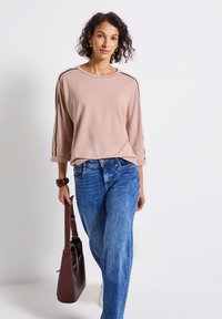 Eine Frau geht, trägt ein hellrosa Langarmshirt, blaue Jeans, weiße Sneaker und hält eine dunkelbraune Handtasche in ihrer rechten Hand.
