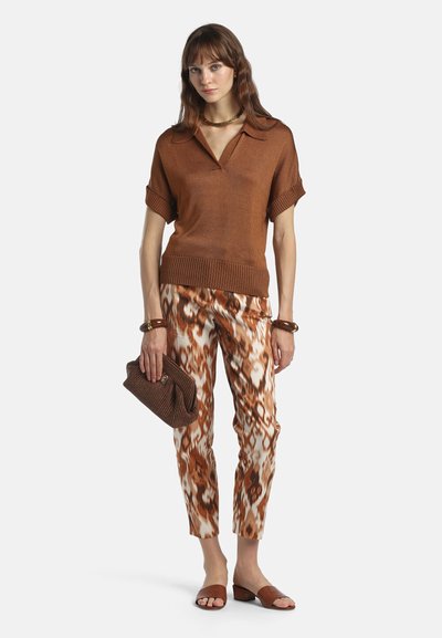 Femme portant un polo marron à manches courtes, un pantalon marron et blanc à motifs, des sandales marron, tenant une pochette marron, avec des bracelets et un collier.