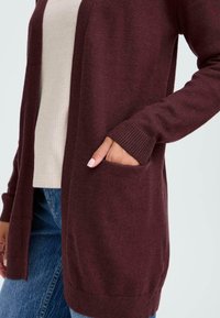 Cardigan lavorato a maglia borgogna con maniche lunghe, apertura frontale, polsini a coste e tasche laterali, indossato sopra un top grigio a coste e jeans blu.