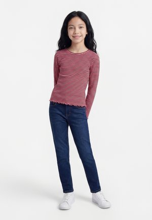 Mlada deklica stoji nasmejana, oblečena v rdeče-črtočasto majico z dolgimi rokavi, modre kavbojke in bele superge na belem ozadju.