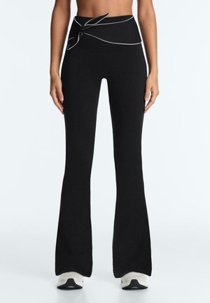 Schwarze, hoch taillierte Schlaghose mit weißen Paspelierungen und einem gebundenen Gürtel, getragen mit weißen Sneakers an einer stehenden Person.