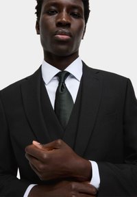 Czarny garnitur w kroju dopasowanym, z białą koszulą i teksturowanym ciemnozielonym krawatem. Wyróżniające detale to slim fit oraz widoczne kieszenie.