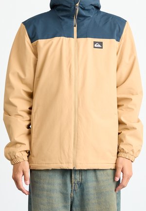 Mann trägt eine beige und dunkelblaue Reißverschlussjacke mit elastischen Bündchen und Jeans, steht vor einem einfarbigen Hintergrund.