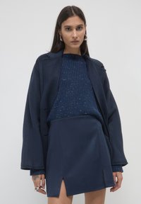 Donna che indossa un cappotto over size blu navy, un maglione a maglia puntinata e una gonna abbinata con spacchi frontali, in piedi davanti a uno sfondo chiaro e neutro.