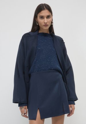 Donna che indossa un cappotto over size blu navy, un maglione a maglia puntinata e una gonna abbinata con spacchi frontali, in piedi davanti a uno sfondo chiaro e neutro.