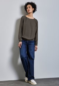 Langärmlige, olivgrüne Bluse mit weitem Hals, kombiniert mit lockeren, dunkelblauen Jeans und weißen Turnschuhen auf einem grauen Hintergrund.