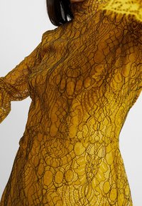 Blusa de encaje amarilla con cuello alto y mangas largas, con intrincados patrones florales negros y un acabado transparente y texturizado.