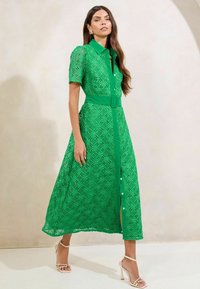 Abito midi verde in pizzo con maniche corte, chiusura con bottoni frontale, vita con cintura e motivi geometrici a taglio. Abbinato a sandali nudi con cinturini.