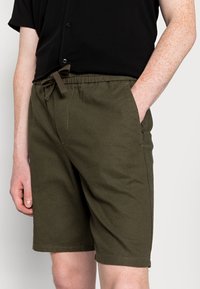 Shorts en coton vert foncé avec une ceinture élastique et un cordon de serrage. Comprend des poches latérales et une coupe décontractée.