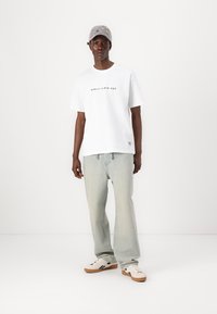 Hvit t-skjorte med svart tekst, lysegrønne jeans og beige sneakers. Grå caps med broderte detaljer fullfører antrekket.