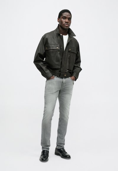 Μαύρο δερμάτινο μπουφάν με δύο τσέπες στο στήθος, πλευρικές τσέπες και κλείσιμο με φερμουάρ. Φοριέται με γκρι τζιν και μαύρα παπούτσια. Μινιμαλιστικός σχεδιασμός και υφή.