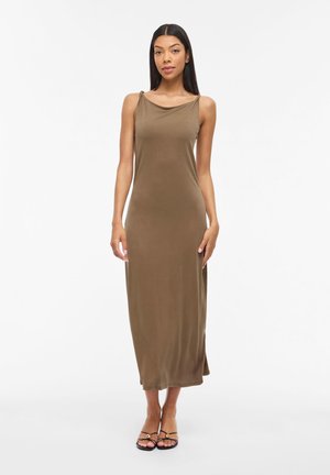 Kobieta stojąca w oliwkowozielonej sukience midi na ramiączkach i czarnych sandałach z odkrytymi palcami na białym tle.