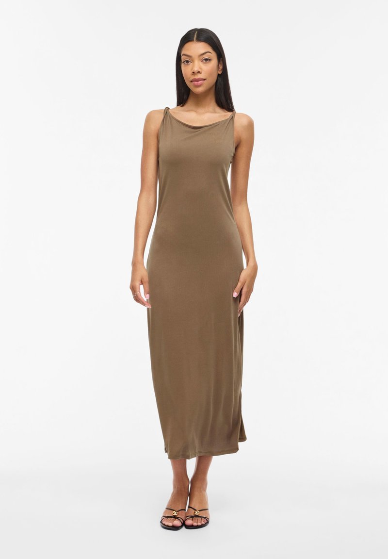 Kvinne står iført en ermeløs midi-kjole i olivengrønt med tynne stropper og svarte sandaler med åpen tå på hvit bakgrunn.