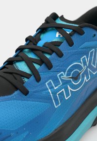 Blauer Sportschuh mit einer strukturierten Mesh-Oberfläche, schwarzen Schnürsenkeln und einer schwarzen Gummisohle, mit dem weißen "HOKA"-Logo an der Seite.