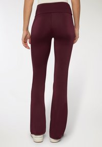 Leggings bordeaux a campana con vita alta, texture liscia e design aderente, che si estendono fino alla caviglia, abbinati a sneakers bianche.
