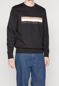 Svart sweatshirt med ett horisontellt randmönster i beige, vitt och mörkgrått över bröstet. Ribbad mudd vid ärmslut och midja.