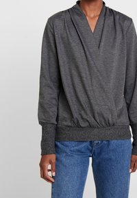 Grijze langarmtop met een V-vormige halslijn, gerimpelde textuur bij de taille en aansluitende mouwen met cuffs, gecombineerd met blauwe spijkerbroek.