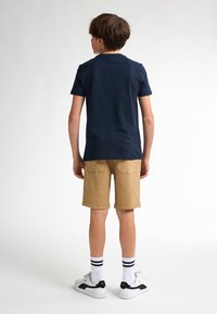 Tricou din bumbac bleu marin, pantaloni scurți khaki și adidași albi cu accente negre. Șosete în dungi. Vedere din spate. O ținută simplă și casual.