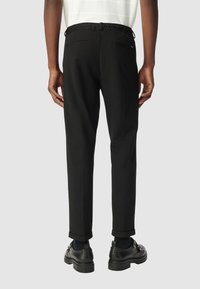 Uomo che indossa pantaloni neri su misura con risvolti, calzini neri e scarpe di pelle nere, mostrato di spalle su uno sfondo grigio uniforme.