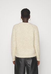 Person mit kurzen lockigen Haaren, die goldene Creolen, einen cremefarbenen Strickpullover und schwarze Lederhosen trägt, vor einem schlichten weißen Hintergrund.