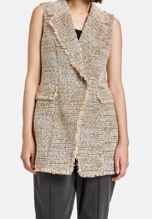 Veste sans manches beige et grise texturée en tweed avec bordure à franges et poches avant à rabat portée sur un haut noir et un pantalon gris.