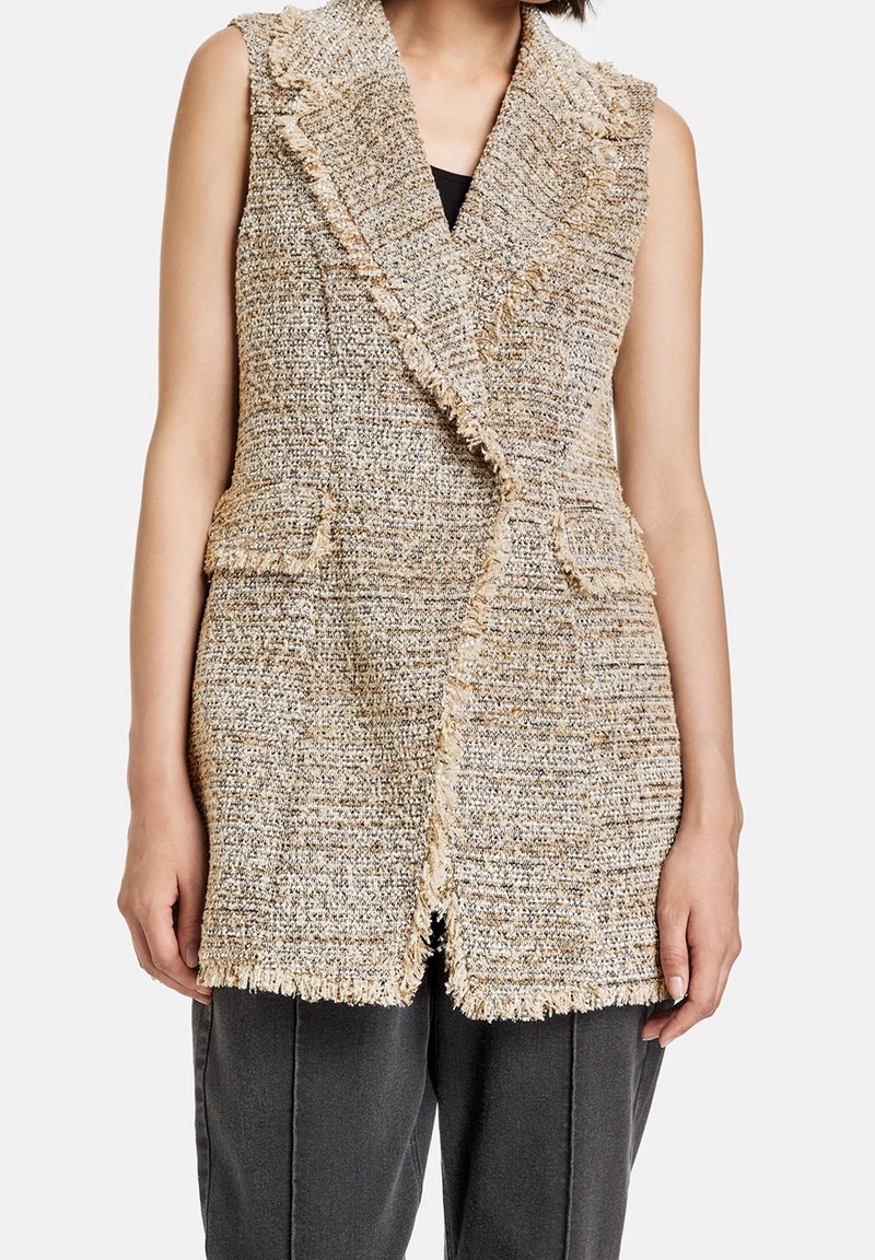 Veste sans manches beige et grise texturée en tweed avec bordure à franges et poches avant à rabat portée sur un haut noir et un pantalon gris.