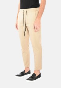 Pantaloni jogger beige con vita elastica, cordino e gambe tapered, abbinati a scarpe slip-on nere con dettagli a nappina.