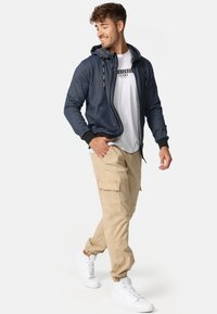 Marinefarbene Zip-Logoshirt mit grauem Futter, schwarze Bündchen; darunter ein weißes T-Shirt. Beigefarbene Cargo-Hosen mit Taschen, kombiniert mit weißen Sneakers.