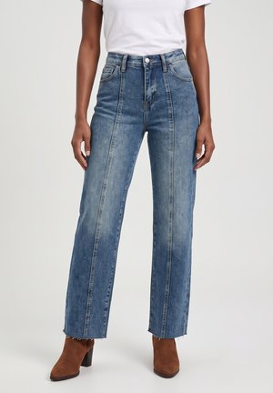 Personne portant un jean bleu taille haute avec des coutures sur le devant et des ourlets bruts, associé à des bottines marron à talons et une chemise blanche à manches courtes.