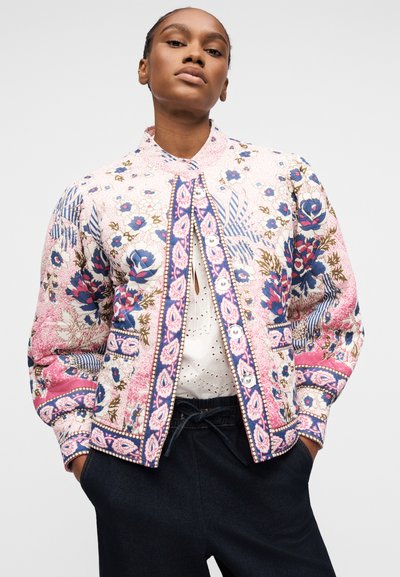 Femme portant une veste à motifs floraux multicolores sur un chemisier blanc en broderie anglaise et un pantalon foncé, debout les mains dans les poches.