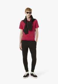 Punainen polo paita mustalla logolla, musta neule solmittuna kaulan ympärille, mustat verkkarit ja ruskeat sneakersit vaaleilla nauhoilla.