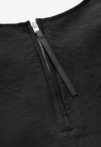 Dettaglio di un indumento nero che presenta una zip metallica visibile e un tessuto testurizzato. Il tirante della zip è nero, con cuciture lungo i bordi.