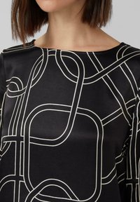 Schwarze Bluse mit einer glatten Textur und einem weißen geometrischen Muster aus überlappenden Linien und Formen. Rundhalsausschnitt.