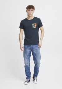 Camiseta navy con un bolsillo de estampado floral y de cebra, llevada con jeans azul claro y zapatillas negras. La tela parece suave y casual.