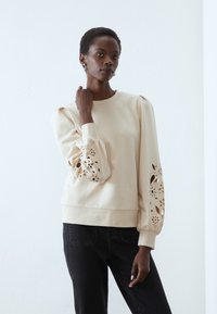 Felpa beige chiaro con maniche a sbuffo, caratterizzata da intricati motivi floreali ritagliati sui polsini. Tessuto morbido, vestibilità morbida.