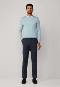 Muž stojící s rukama v kapsách, oblečený v světle modrém svetru, námořnických kalhotách a tmavých botách na jednobarevném pozadí.