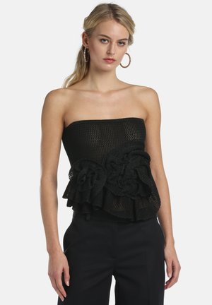 Moteris dėvi juodą be petnešėlių megztą palaidinę su didelėmis raukšlėtomis gėlių detalėmis, derinama su juodomis kelnėmis ir didelėmis lankelio formos auskarais.