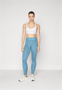 Bjela sportska brusnica s podesivim trakama, u kombinaciji s visokim svijetloplavim tajicama s "PRO" logom i malim bijelim Nike oznakom.