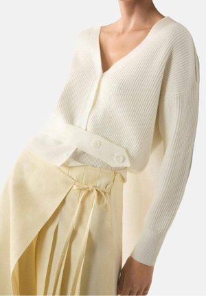 Vrouw draagt een crèmekleurige geribbelde cardigan met knoopdetails en een lichtgele wikkelrok die in de taille is gestrikt.