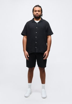 Mand med fletninger iført en sort kortærmet skjorte med knapper og matchende sorte shorts, hvide sokker og hvide sneakers, stående.