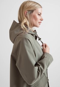 Water-resistant green rain jacket with a hood, adjustable drawstring, and snap-button cuffs. Features a smooth texture and minimalist design.