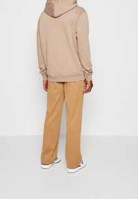 Person som bär beige huvtröja och kamelfärgade vida byxor, stående med ryggen mot kameran mot en enfärgad vit bakgrund.