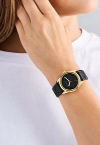Kullatud tooni käekell musta tekstuuriga kellapinnaga, ümar korpus ja must nahkrihm. Omab minimalistlikke tunniindikaatoreid ja lihtsalt kroonit.