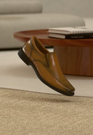 Brązowy, skórzany elegancki but wsuwany unoszący się nad beżowym, teksturowanym dywanem z drewnianym stołem i sofą w tle.