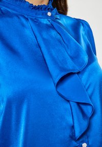 Blusa in satin blu con colletto a volant, pieghe e un dettaglio di bottone decorativo sul lato. Texture liscia e lucente con un colore vivace.