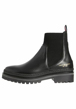 Botas Chelsea negras de cuero texturizado, con paneles laterales elásticos, suelas resistentes y un acento de logo dorado en el lateral.