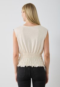 Haut beige clair sans manches avec détail de taille smockée et ourlet volanté ; texture lisse. Porté avec un jean noir taille haute.